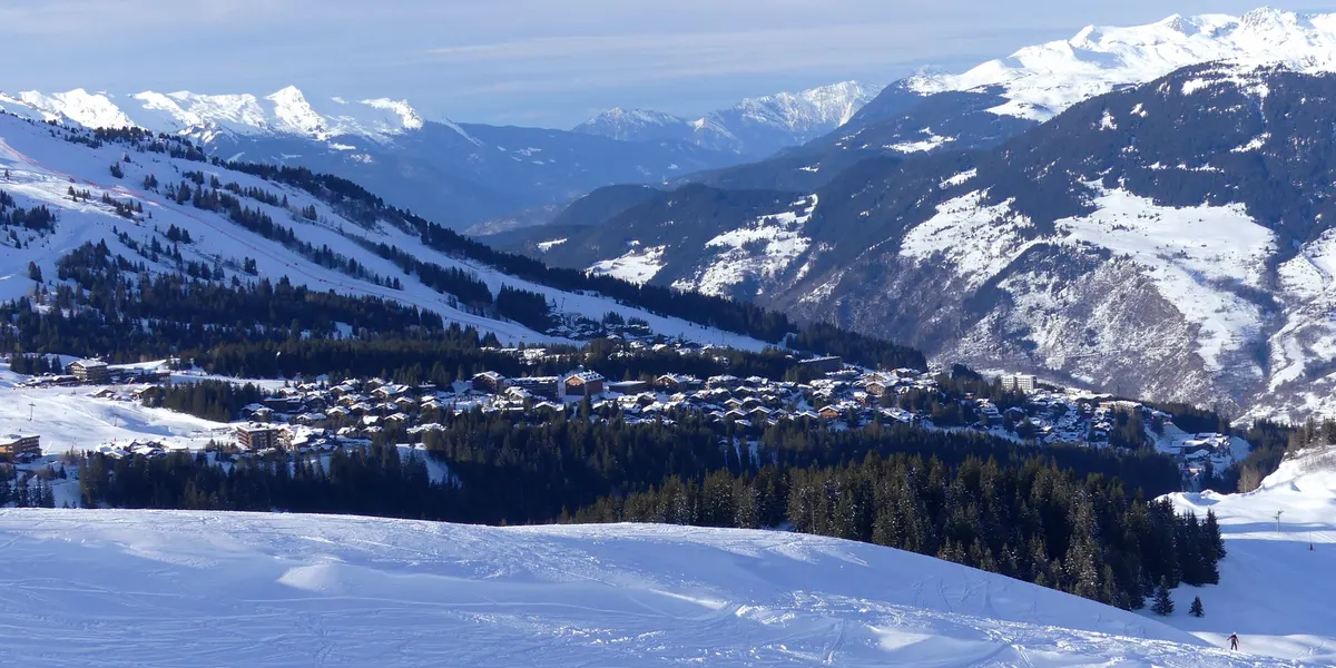 Chalets of Courchevel among pine forest below a groomed piste, with snowy mountains behind