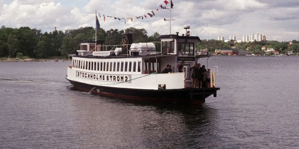 A white Stockholm archipelago passenger boat with passengers on deck, flags flying, wooded islands behind