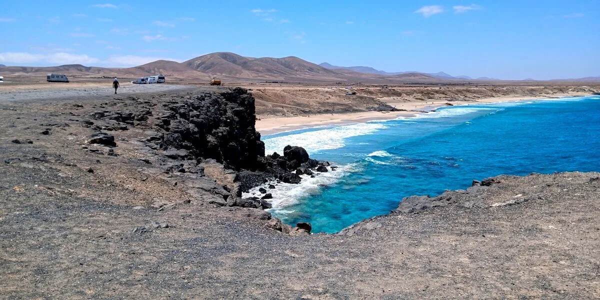 A dark volcanic headland sheltering a turquoise bay at El Cotillo on Fuerteventura, with bare winter hills behind